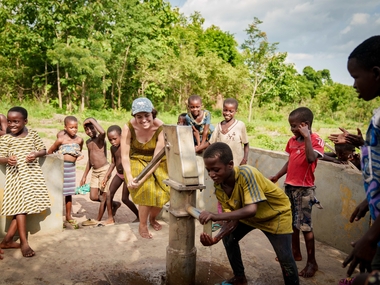 Eine Gruppe von Kindern steht mit einer erwachsenen Person an einer handbetriebenen Wasserpumpe im Freien. Die erwachsene Person betätigt die Pumpe, während ein Kind Wasser in einen Behälter füllt. Die anderen Kinder schauen lächelnd zu. Im Hintergrund stehen grüne Bäume, die Szene wirkt lebendig und gemeinschaftlich.